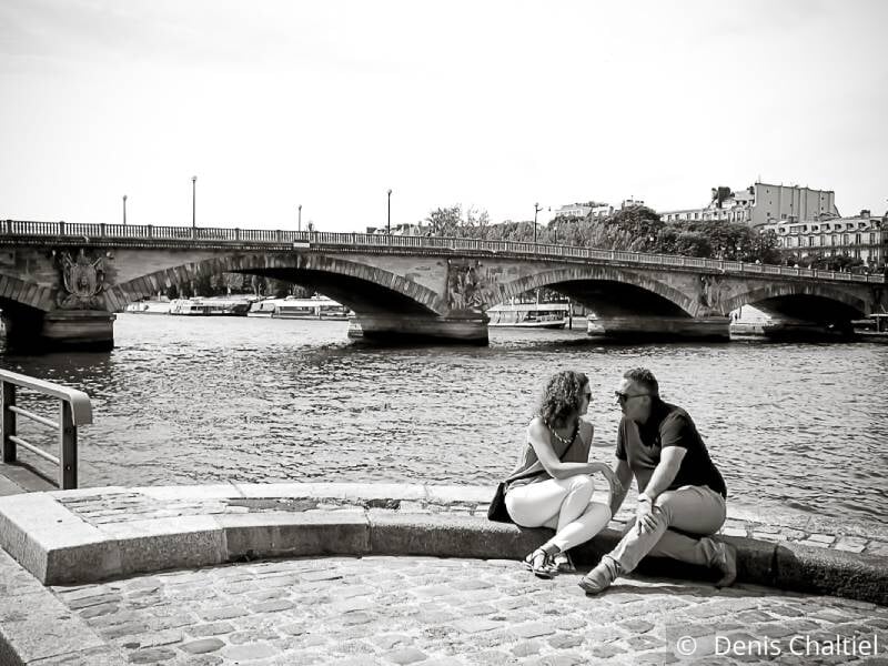 Un couple est assis au bord de l'eau et se regarde amoureusement. Photo en noir et blanc avec un pont en arrière plan.